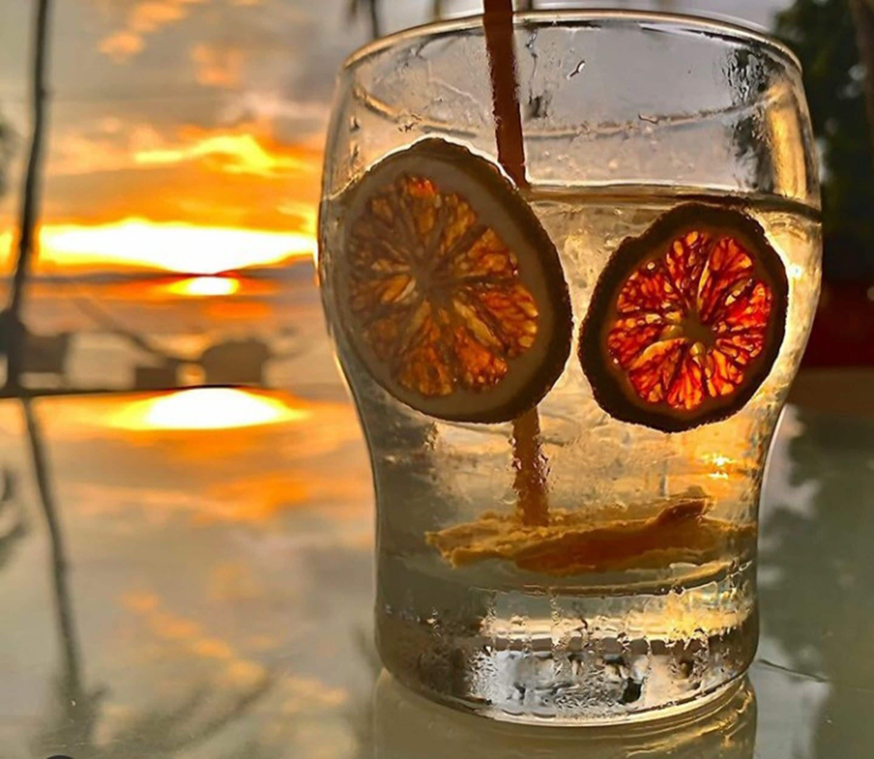 A glass with dried fruit slices and a straw, reflecting a sunset.