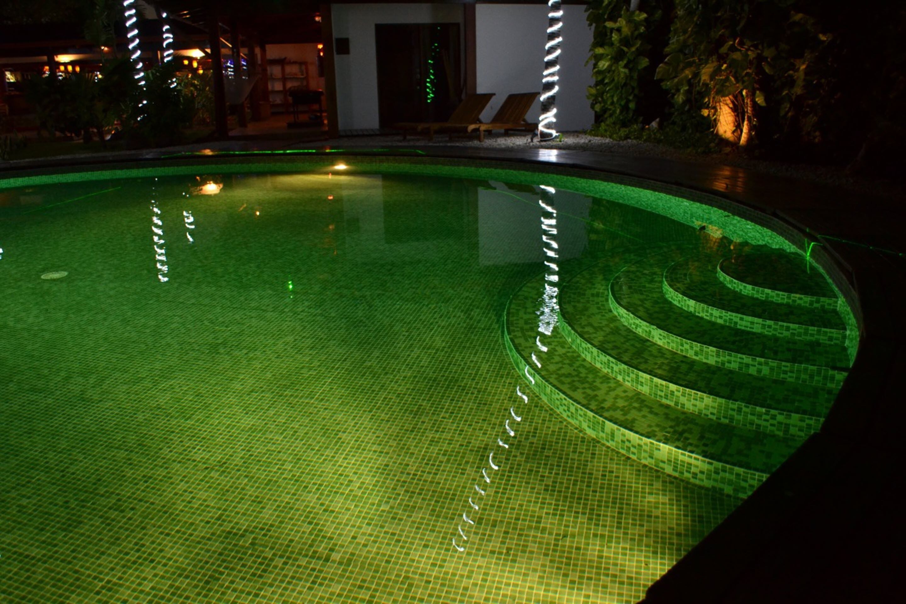 Vista nocturna de una piscina con diseño espiral, rodeada de un área ajardinada con árboles y un edificio al fondo.