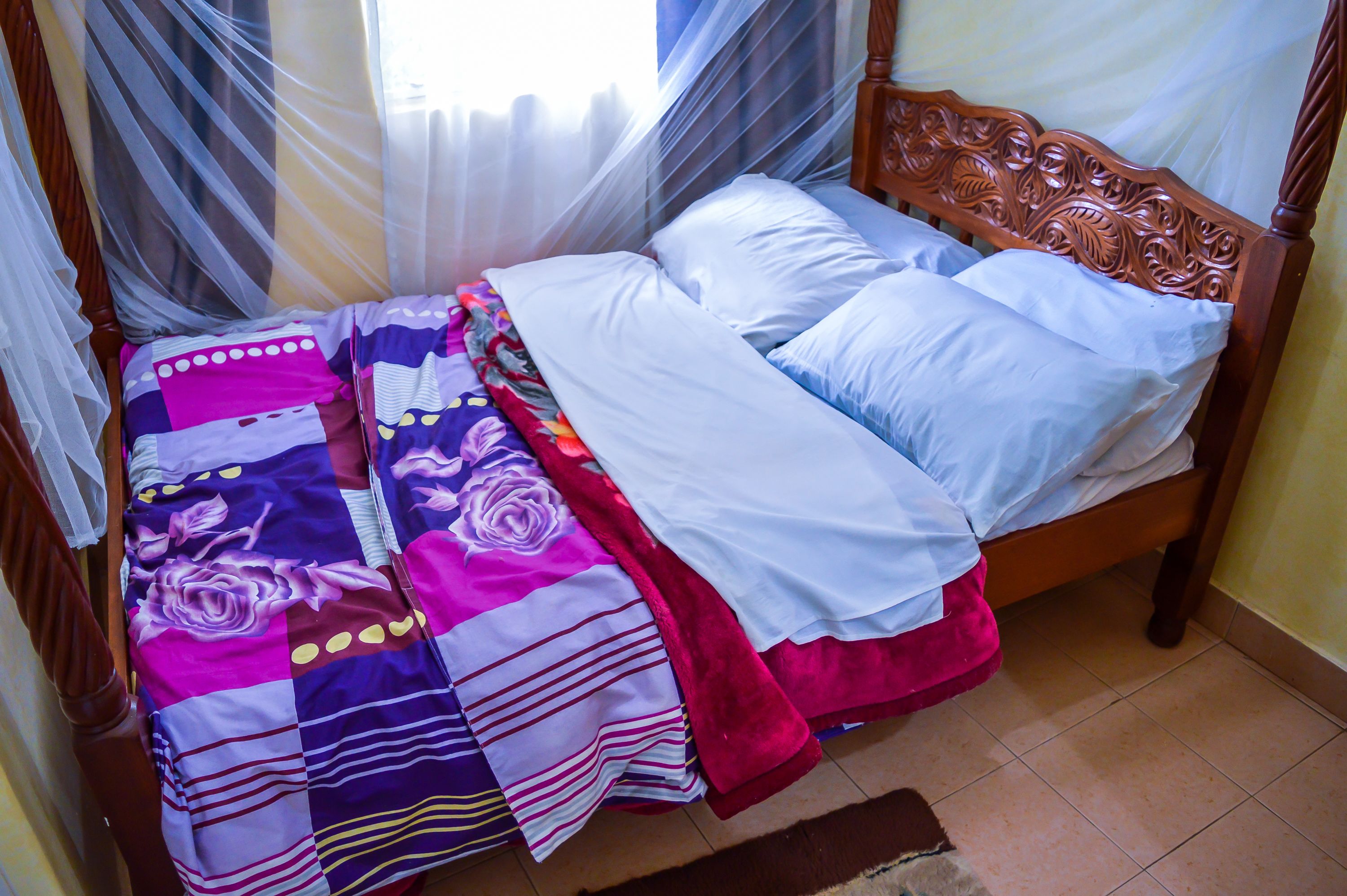 A bed with a colorful quilt, a wooden headboard, and sheer curtains.