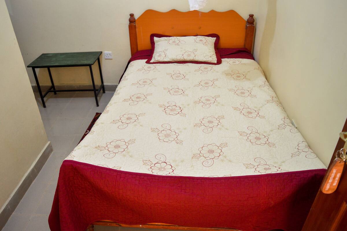 A small, neatly made bed with a floral-patterned bedspread and a red blanket. There is a wooden headboard and a small table beside the bed.