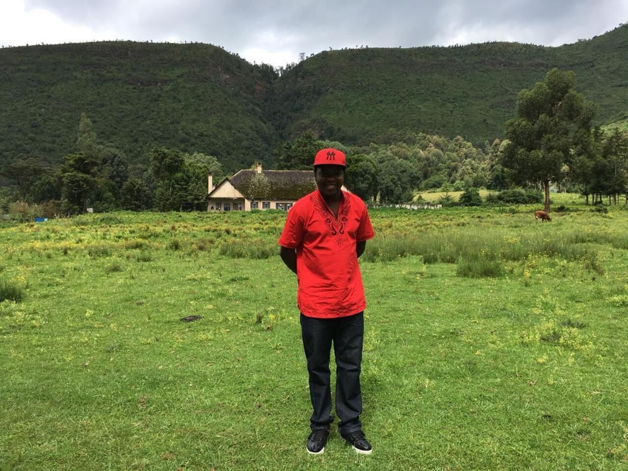 A person standing in a grassy field with a house in the background and hills in the distance.
