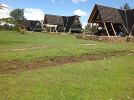 Una hilera de cabañas tipo A con cimientos de piedra y vigas de madera, ubicadas en un campo cubierto de hierba con árboles al fondo.