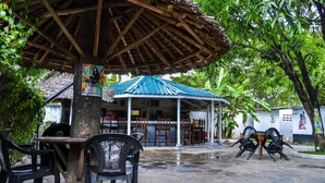 An outdoor seating area with a thatched roof and a covered pavilion. Tables and chairs are arranged around a central tree. - Sanana Conference Center and Holiday Resort (Mombasa)