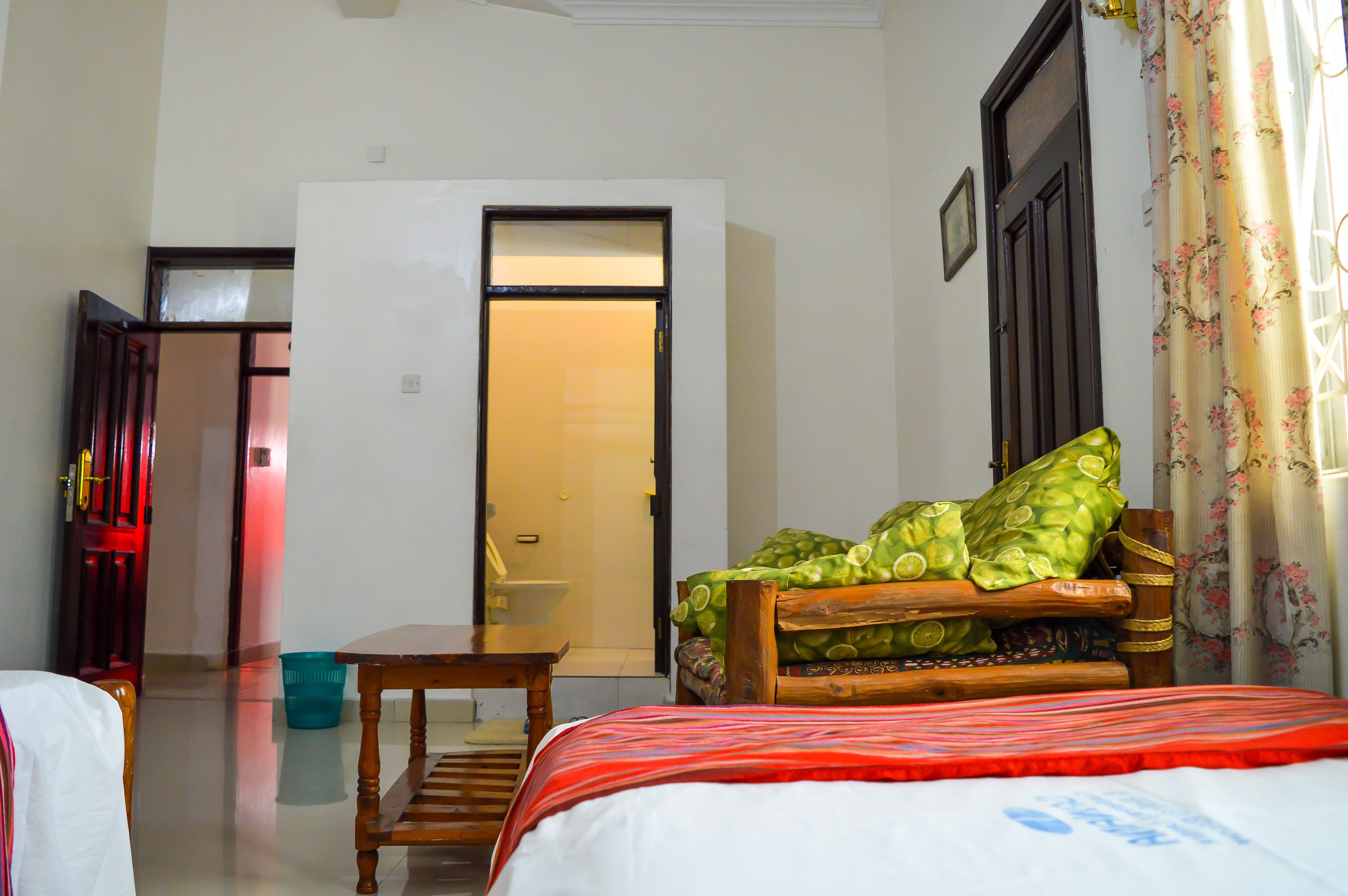 Habitación de hotel con una cama, un banco de madera, una mesita y una puerta que da a un baño.