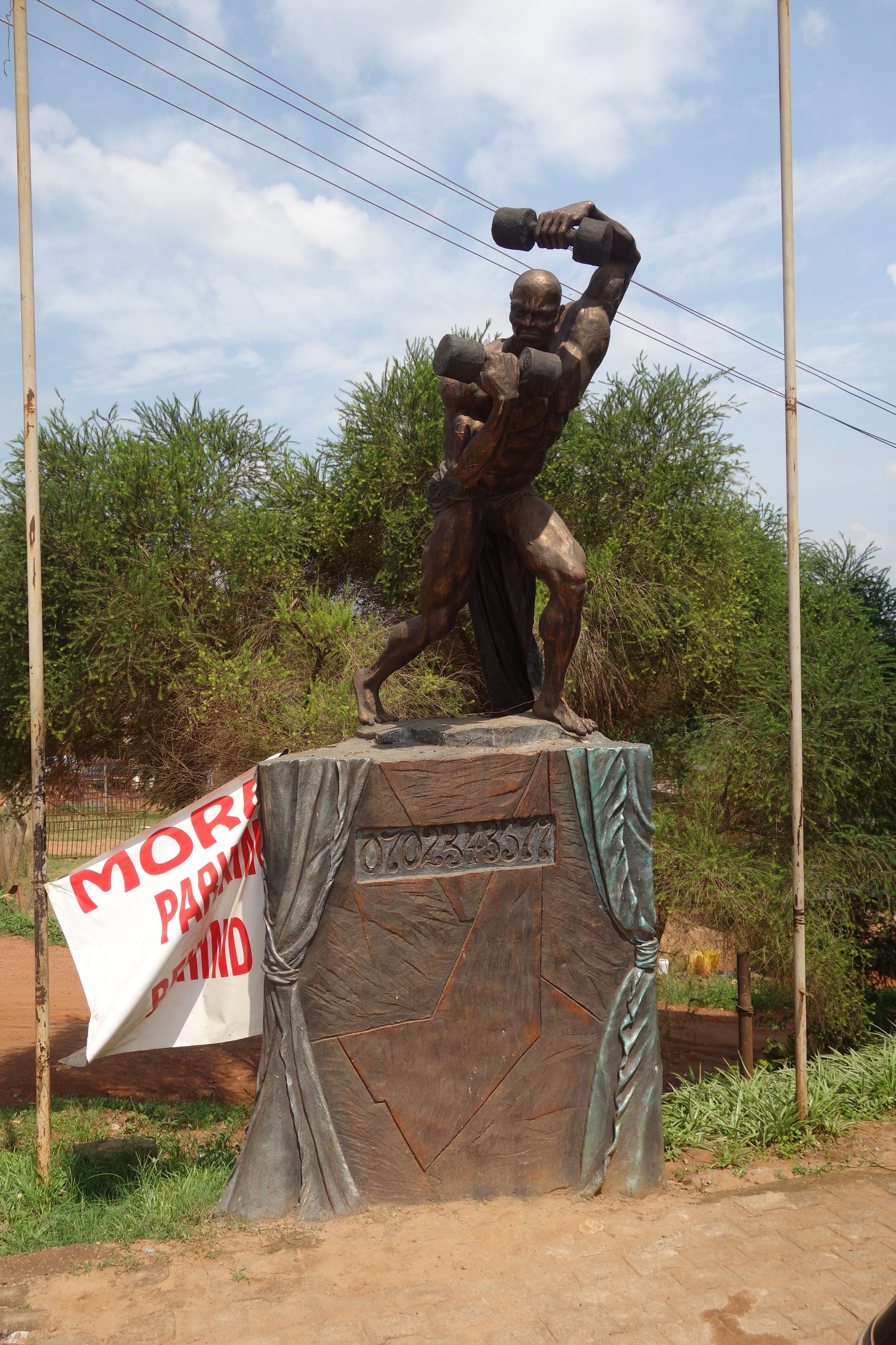 Una estatua de bronce de un hombre levantando pesas, montada sobre un pedestal con un cartel que dice 