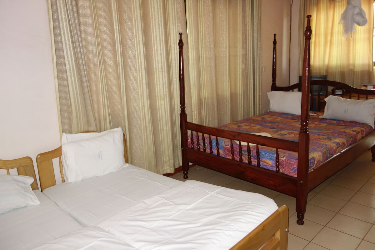 A double bed with a wooden frame and a colorful bedspread, a single bed with a wooden frame and white bedding, and a wooden nightstand with a lamp.