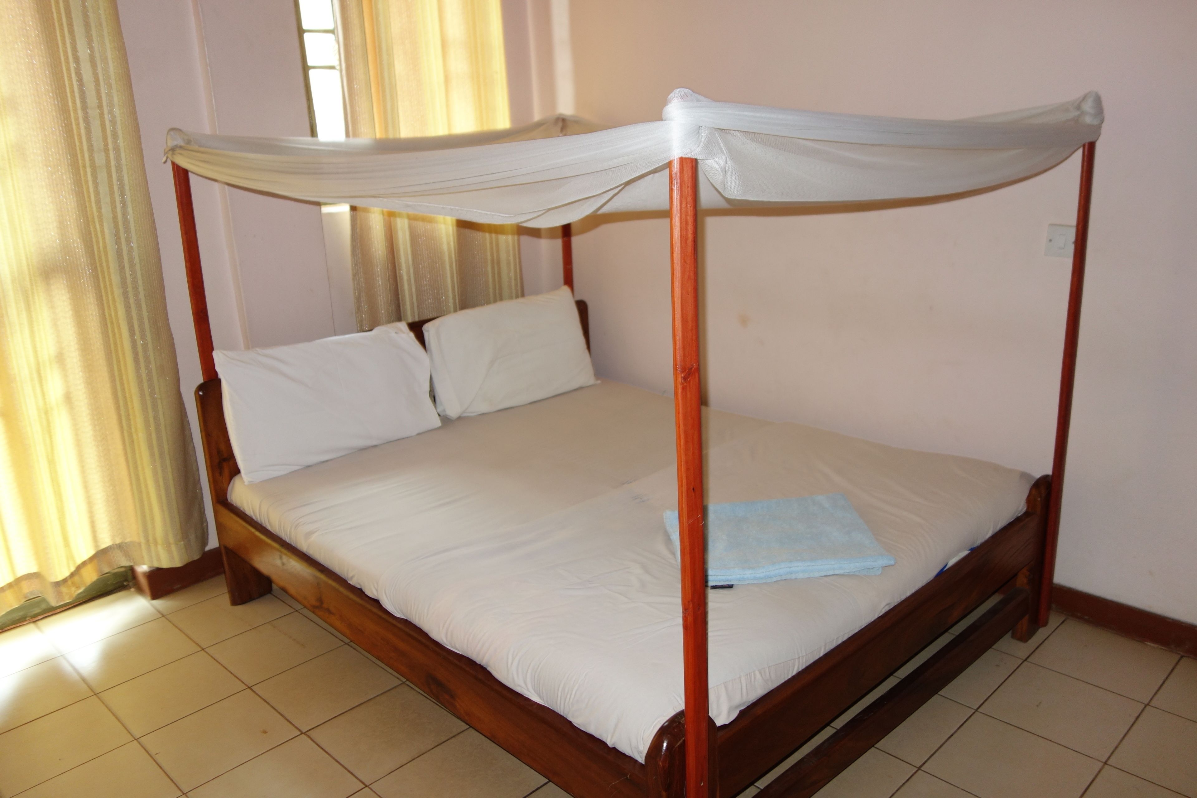 A four-poster bed with white linens and a blue towel, wooden frame, and a window with curtains.