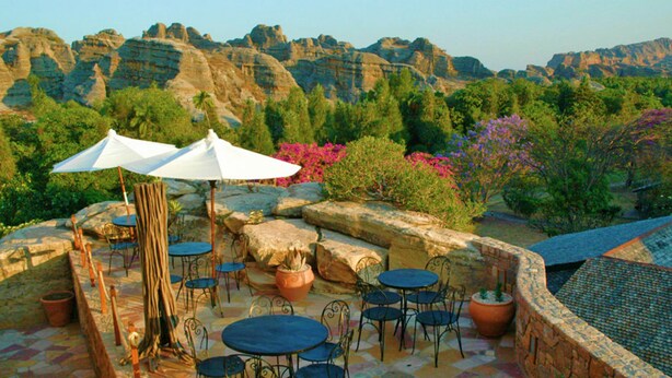 Un patio al aire libre con suelo de piedra, mesas redondas y sillas, rodeado de follaje y formaciones rocosas.