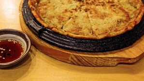 Una tortita redonda y dorada sobre una tabla de madera, con un recipiente pequeño de salsa roja al lado.