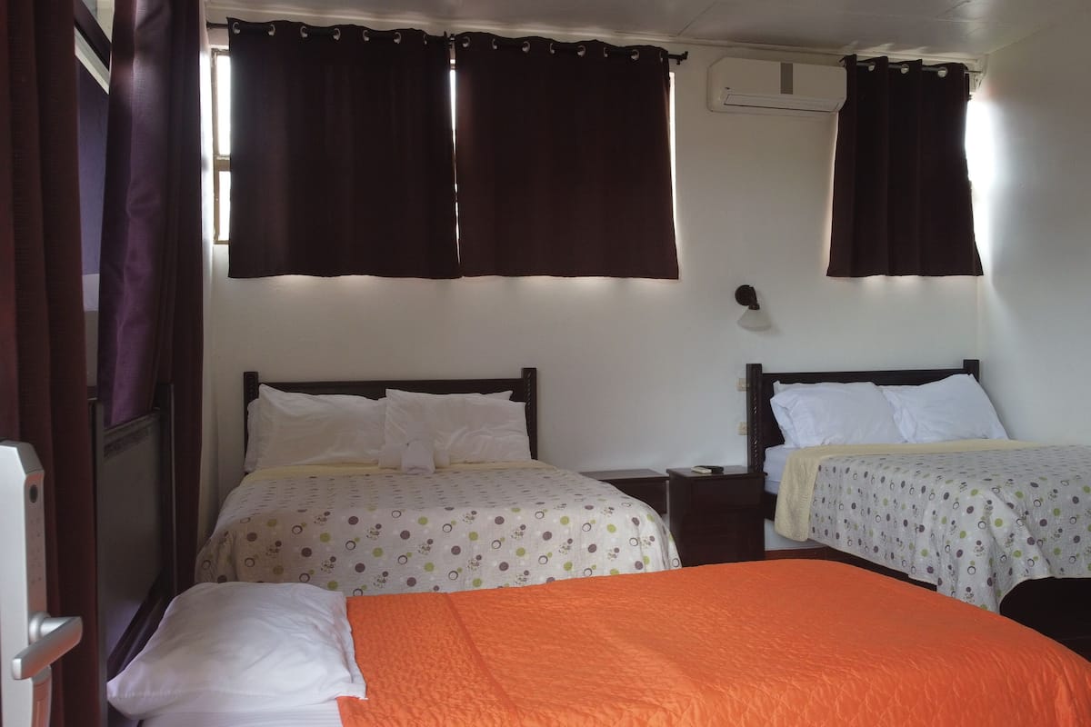 A hotel room with two beds, each with a different patterned bedspread, a nightstand, a wall-mounted lamp, and an air conditioning unit.
