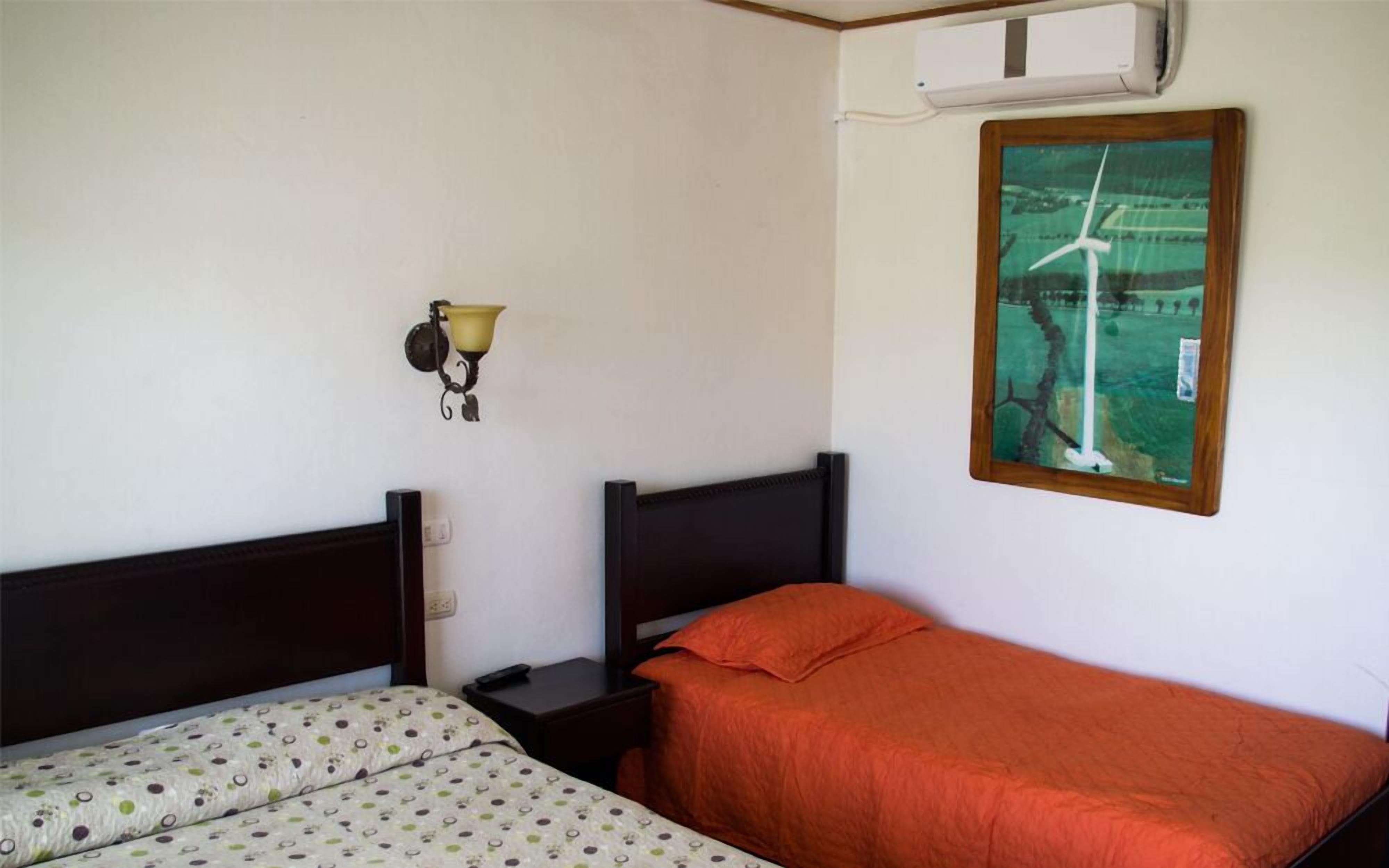 Une chambre avec deux lits, un climatiseur fixé au mur, une lampe et un tableau représentant une éolienne.