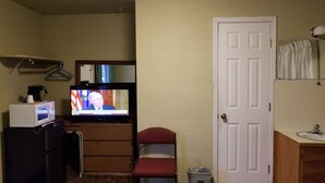 Une petite chambre d’hôtel avec un téléviseur diffusant un programme d’information, une commode en bois, une chaise rouge, une porte blanche et un petit coin lavabo.