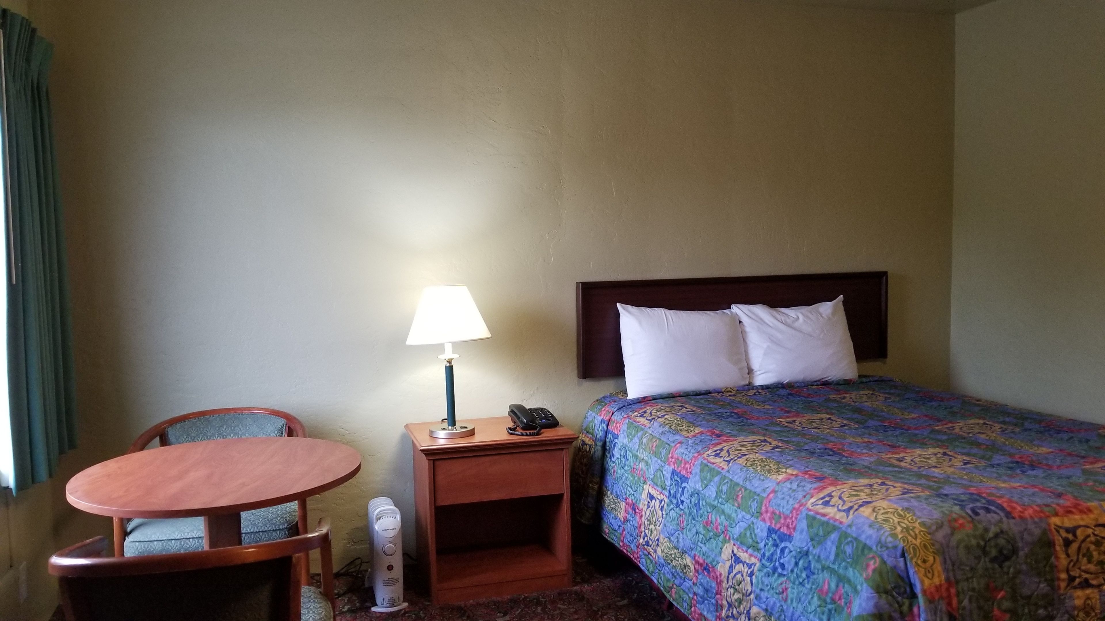 Quarto de hotel com cama, mesa de cabeceira com abajur, uma cadeira e uma pequena mesa.