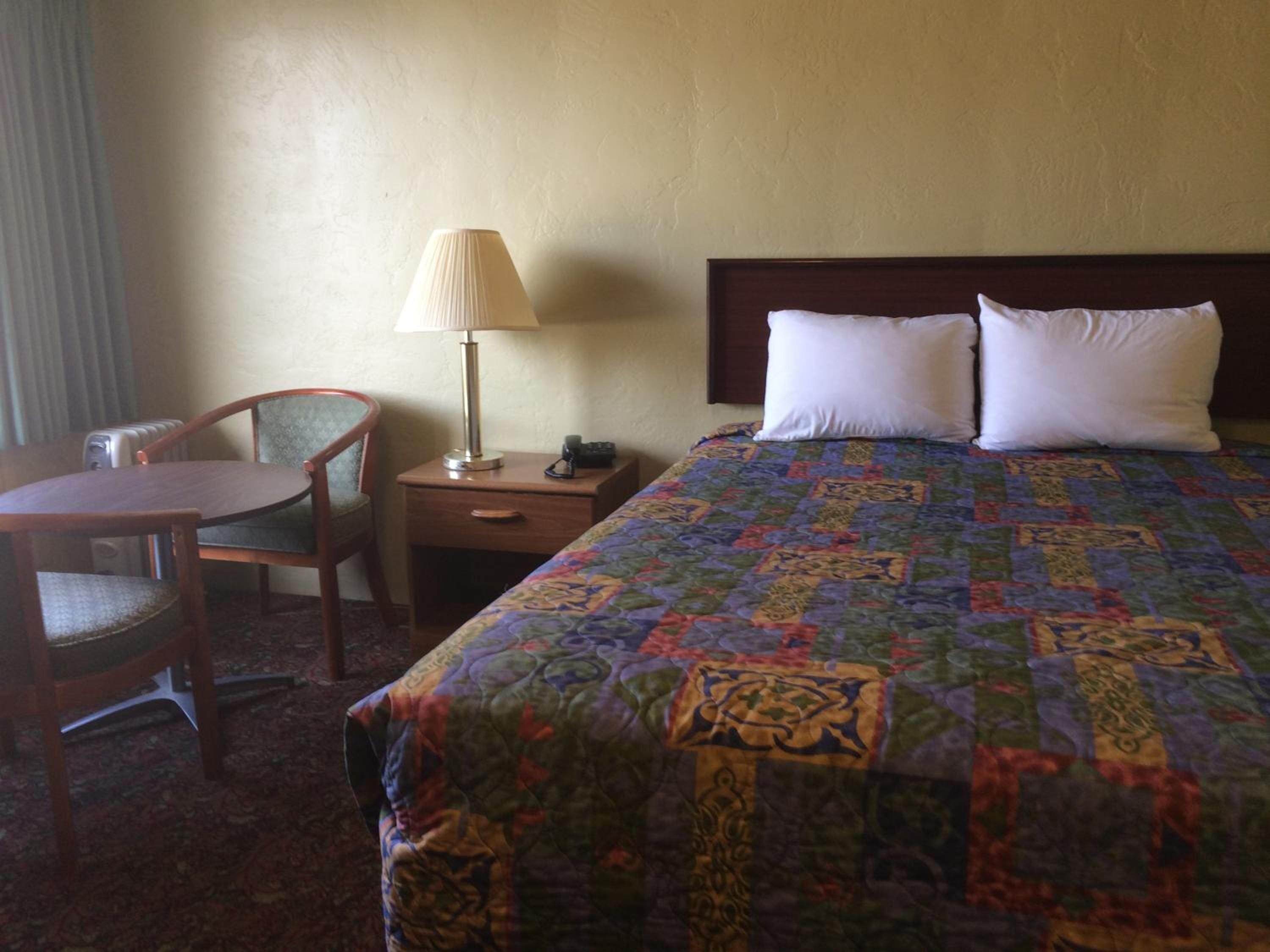 Habitación de hotel con una cama, dos almohadas, una mesita de noche con lámpara, una silla y una pequeña mesa.