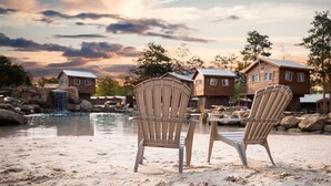 Two Adirondack chairs on a sandy beach with a view of wooden cabins and a small waterfall in the background.