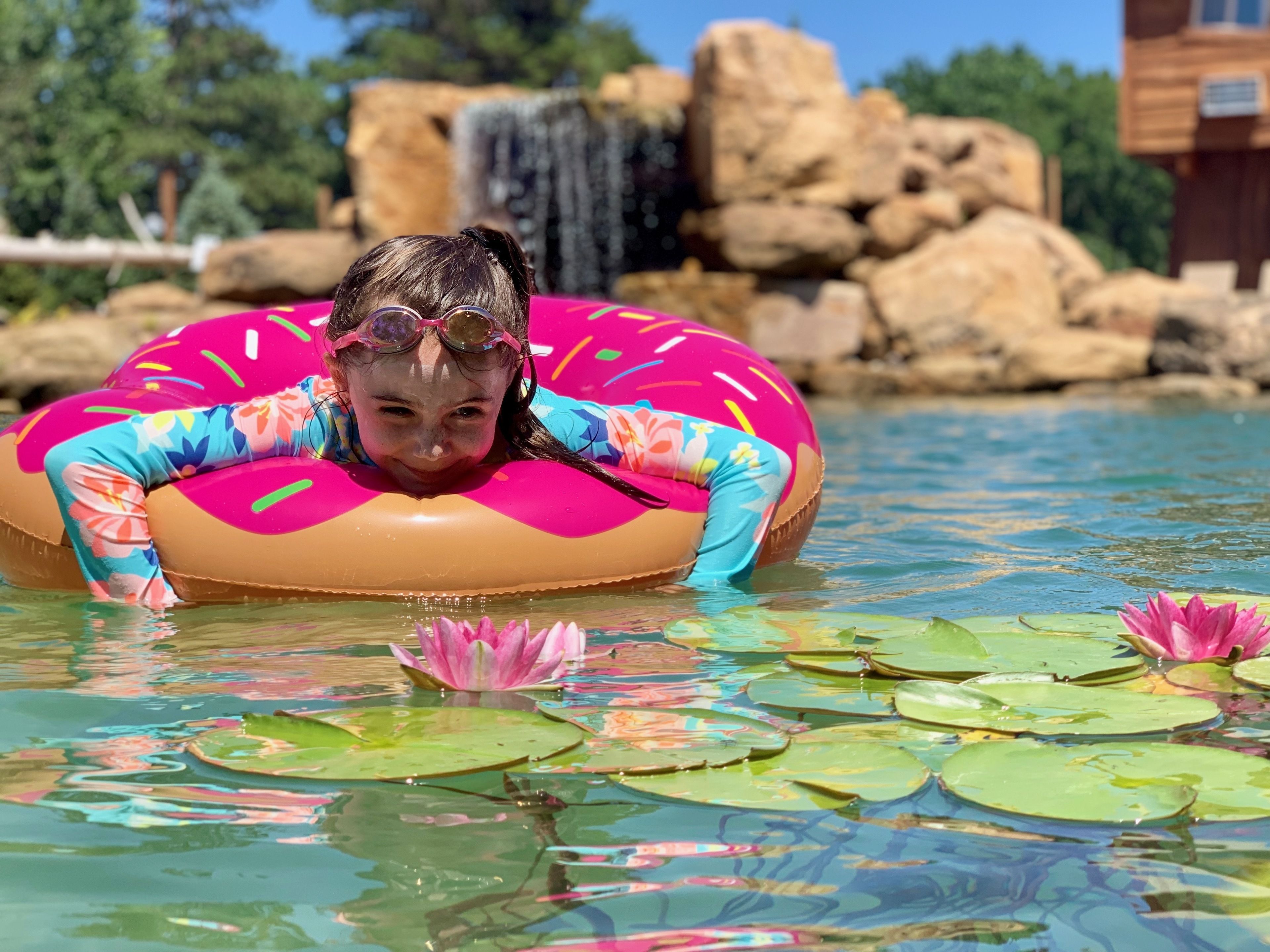 Una persona en una piscina con un flotador rosa, rodeada de nenúfares y una cascada al fondo.