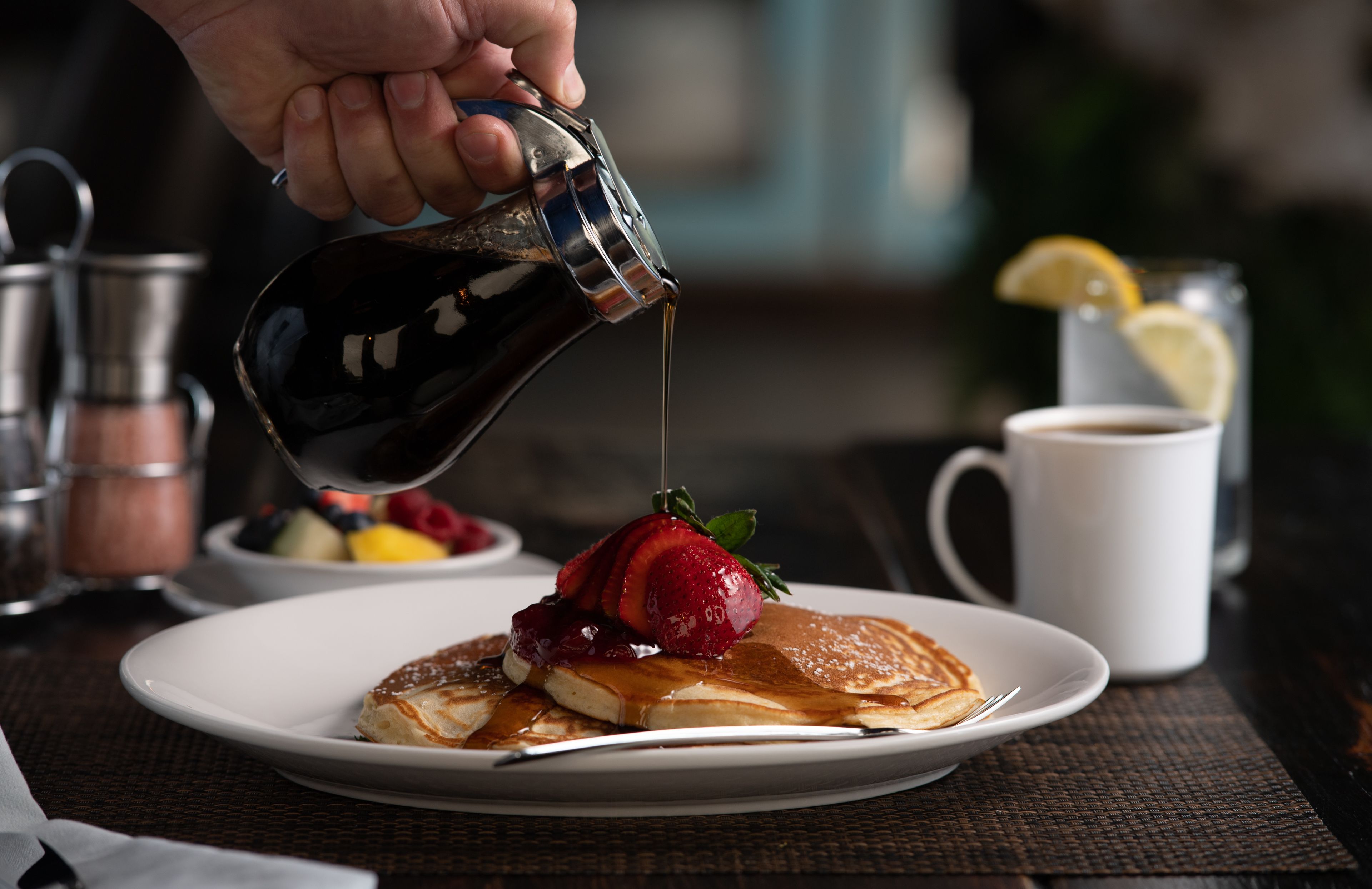 En hand häller sirap över pannkakor som ligger på en tallrik tillsammans med jordgubbar.