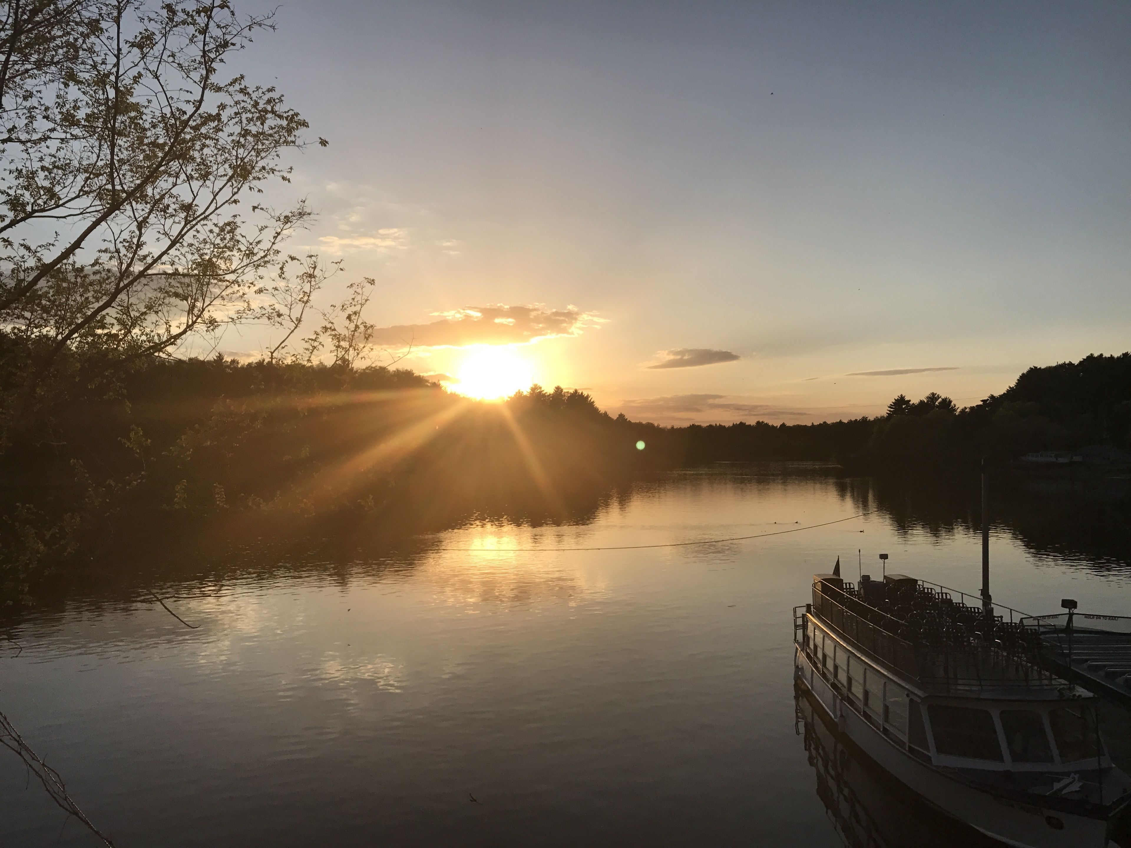 A calm river with a boat docked on the right. Sunlight is reflecting off the water, and there are trees along the riverbank.