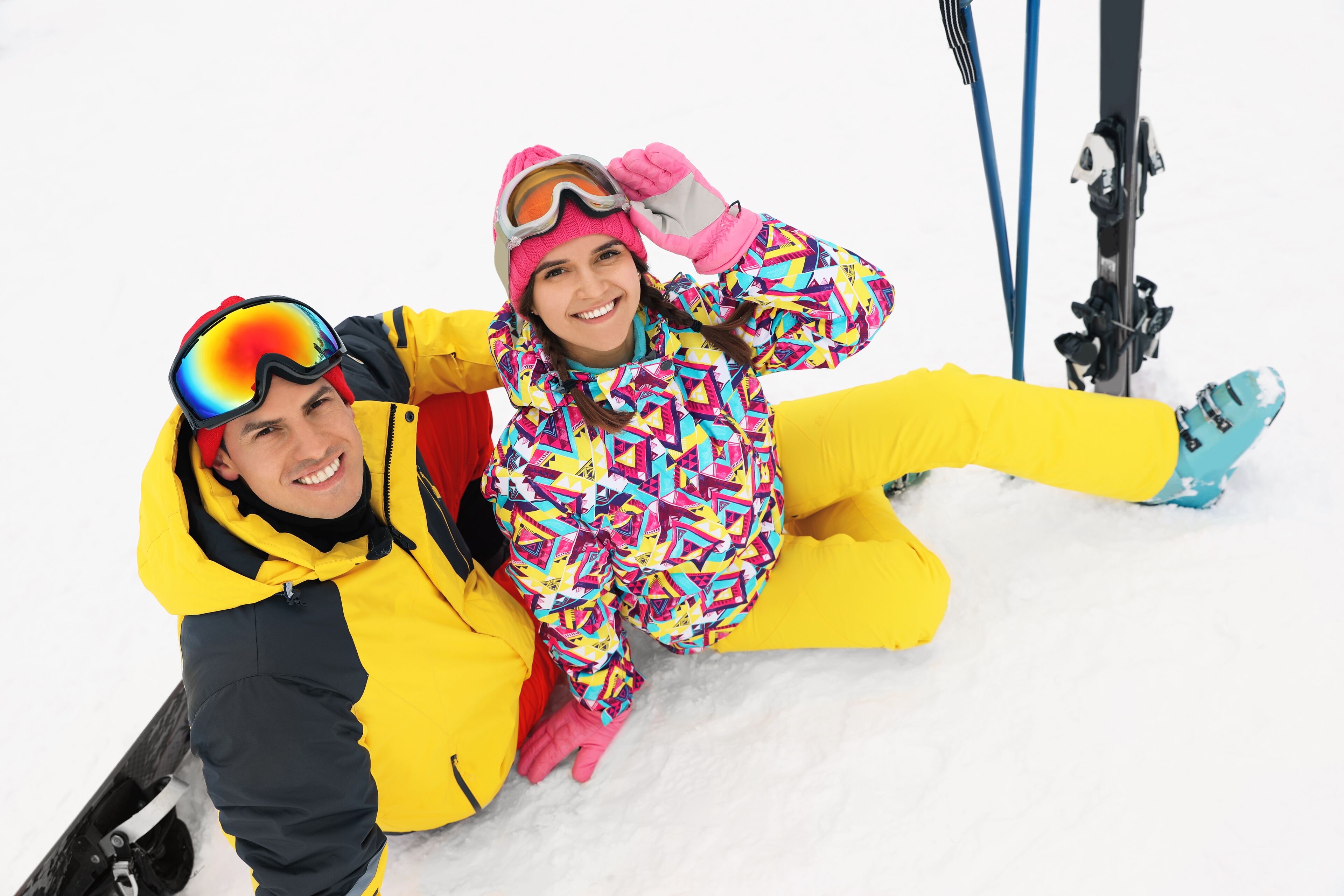 Two people sitting on snow with skis nearby.