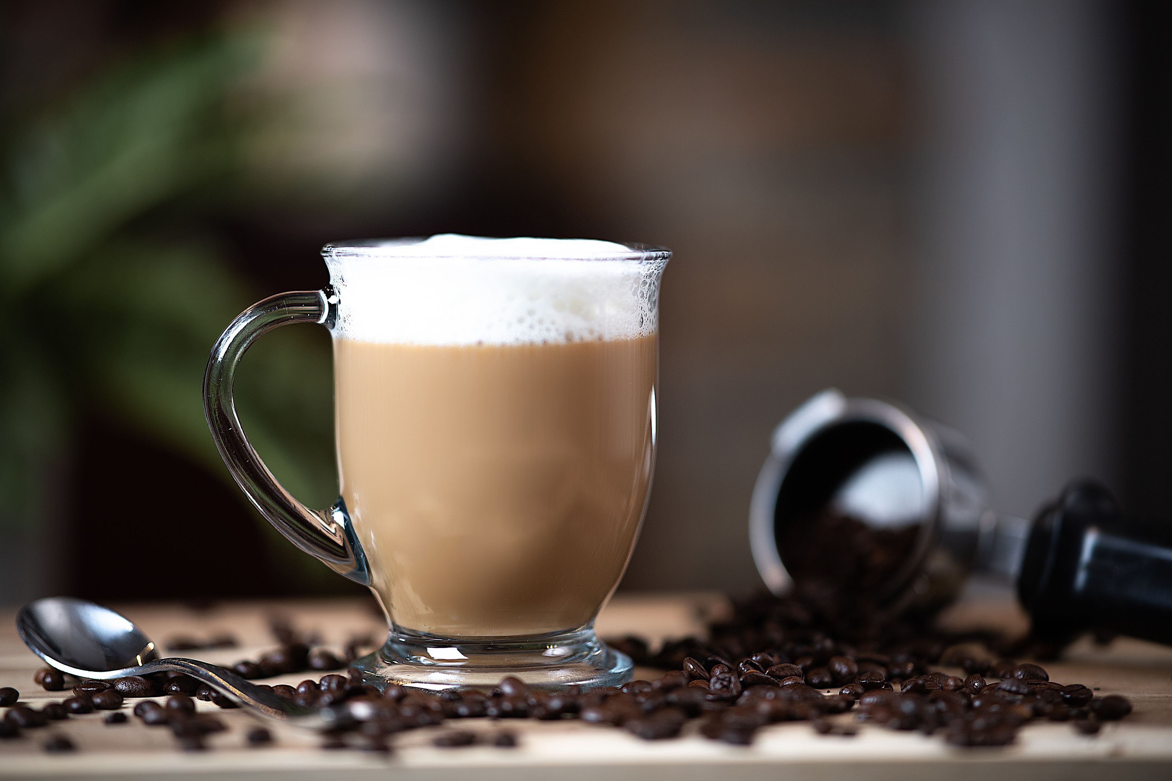 Una taza de vidrio transparente llena de una bebida espumosa, una cuchara y granos de café esparcidos sobre una superficie de madera.