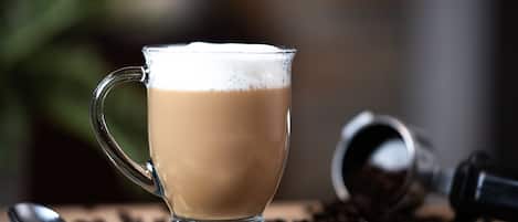 Una taza de vidrio transparente llena de una bebida espumosa, una cuchara y granos de café esparcidos sobre una superficie de madera.