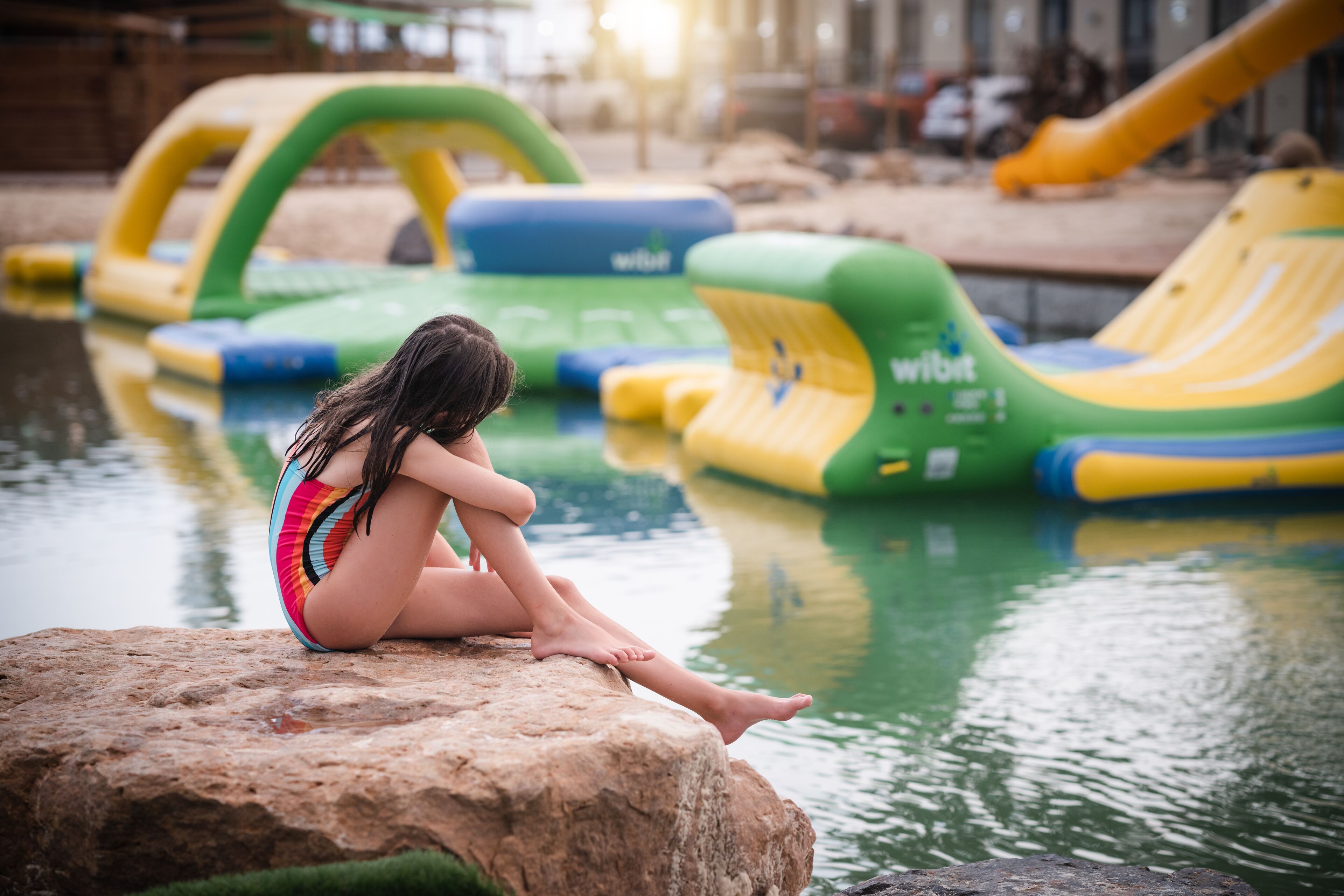 A child sitting on a rock by a water body with inflatable play structures in the background.
