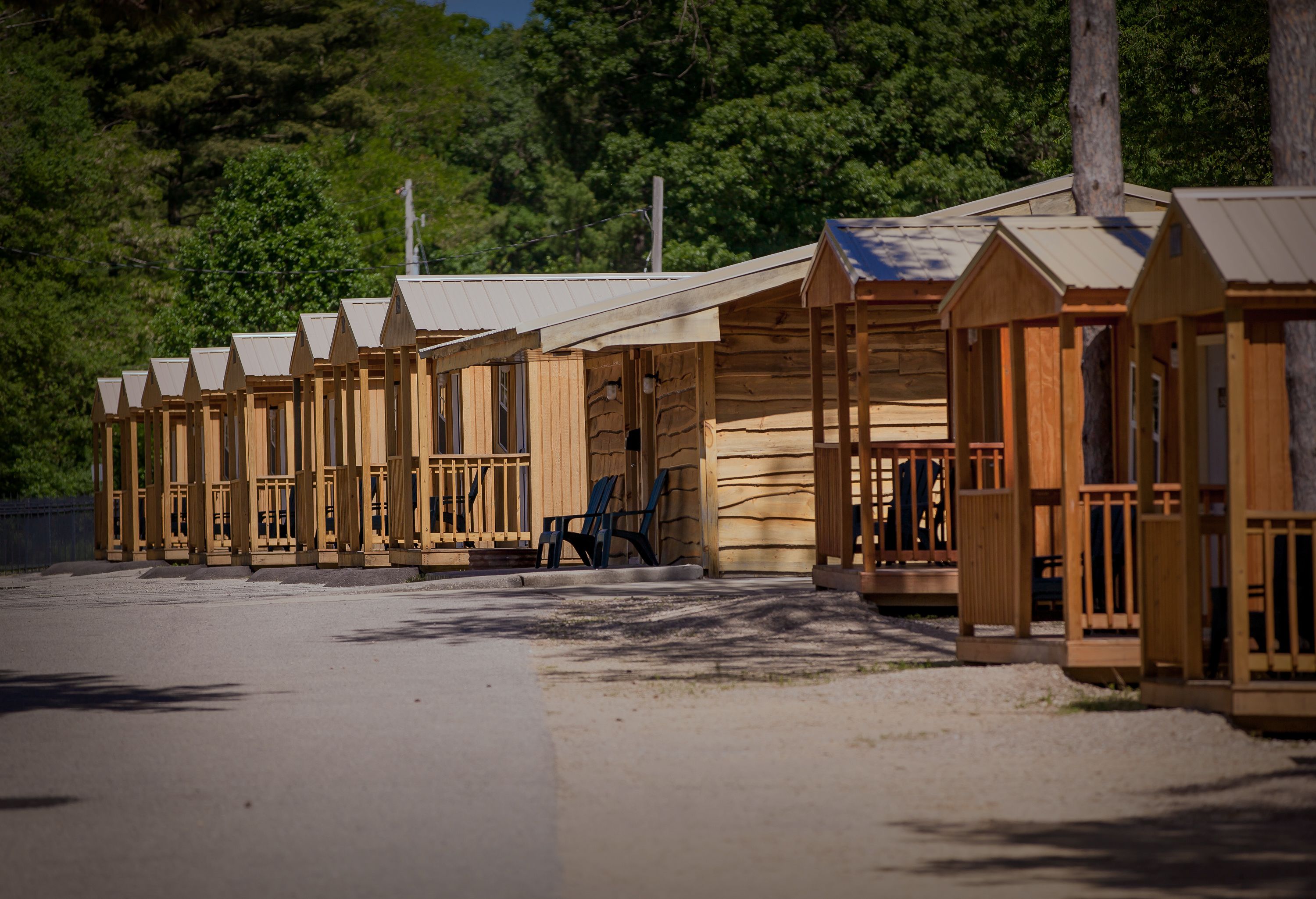 A row of wooden cabins with pitched roofs, arranged along a gravel path with a forested backdrop.