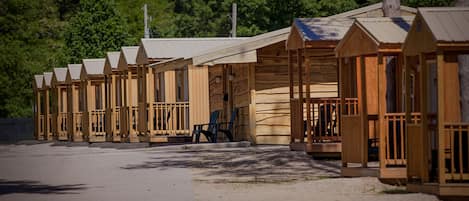 A row of wooden cabins with pitched roofs, arranged along a gravel path with a forested backdrop.