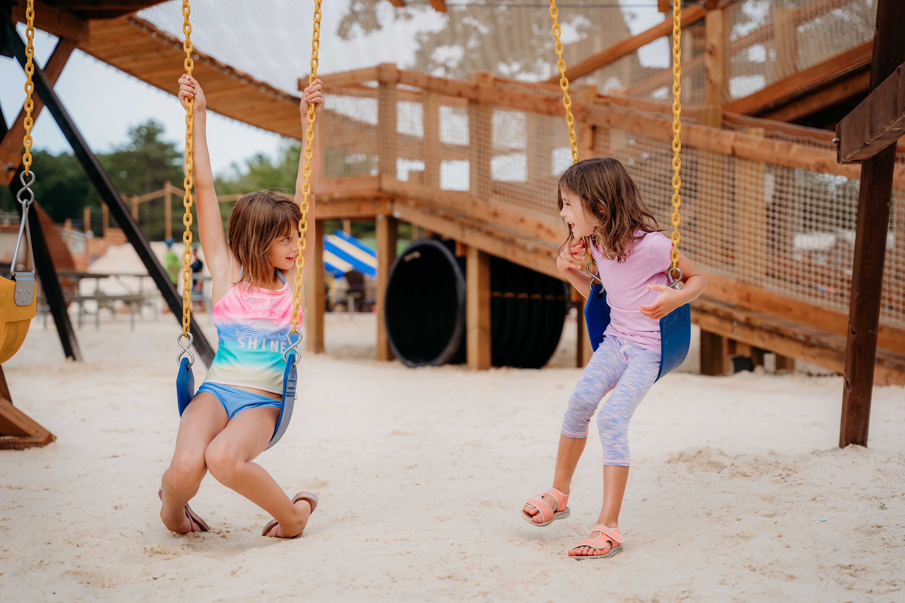 Two children on swings at a playground with a wooden structure and netted safety barrier.