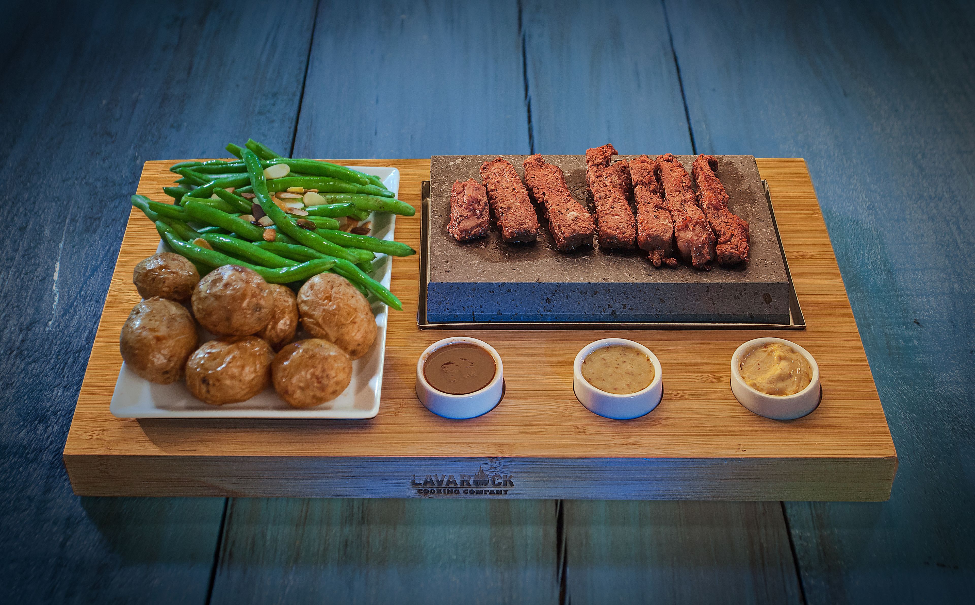 A wooden serving board with green beans, meat, and small round items, accompanied by three small bowls of sauces.