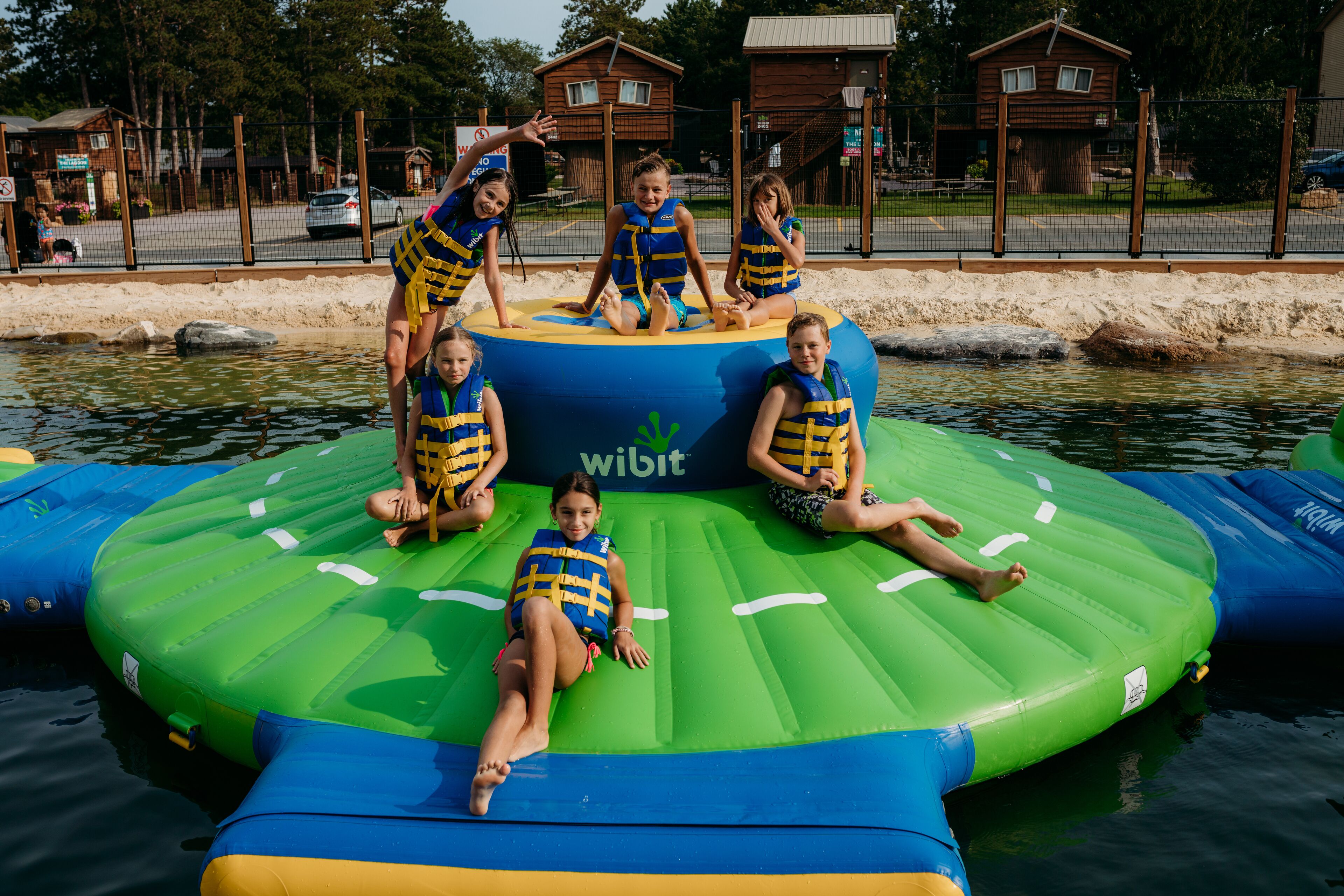 A group of children on an inflatable raft with a 'Wibit' logo, surrounded by water and a fenced area with buildings in the background.