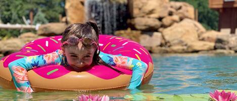 Una persona en una piscina con un flotador rosa, rodeada de nenúfares y una cascada al fondo.
