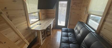 A compact cabin with a wooden interior, a black leather sofa, a small table, and a TV mounted on the wall.