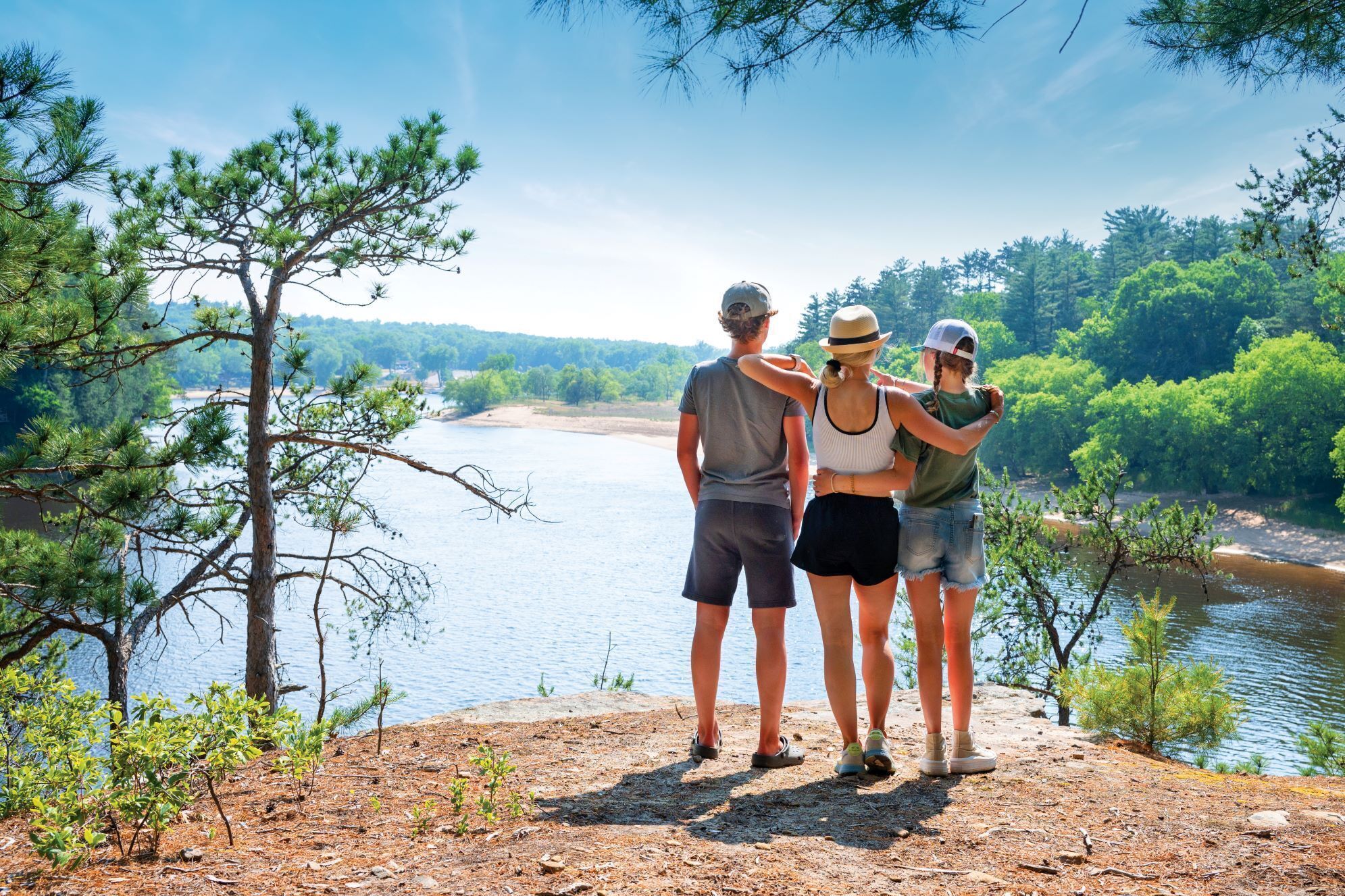 Three people standing on a dirt path overlooking a river, surrounded by trees and greenery.