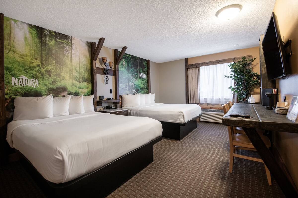 A hotel room with two beds, a desk, a TV, and a large forest-themed wall mural.