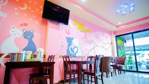 A restaurant interior with pink walls, a flat-screen TV, wooden tables and chairs, and a view of the street through large windows. - Sweet Dreams Hometel (Krabi)