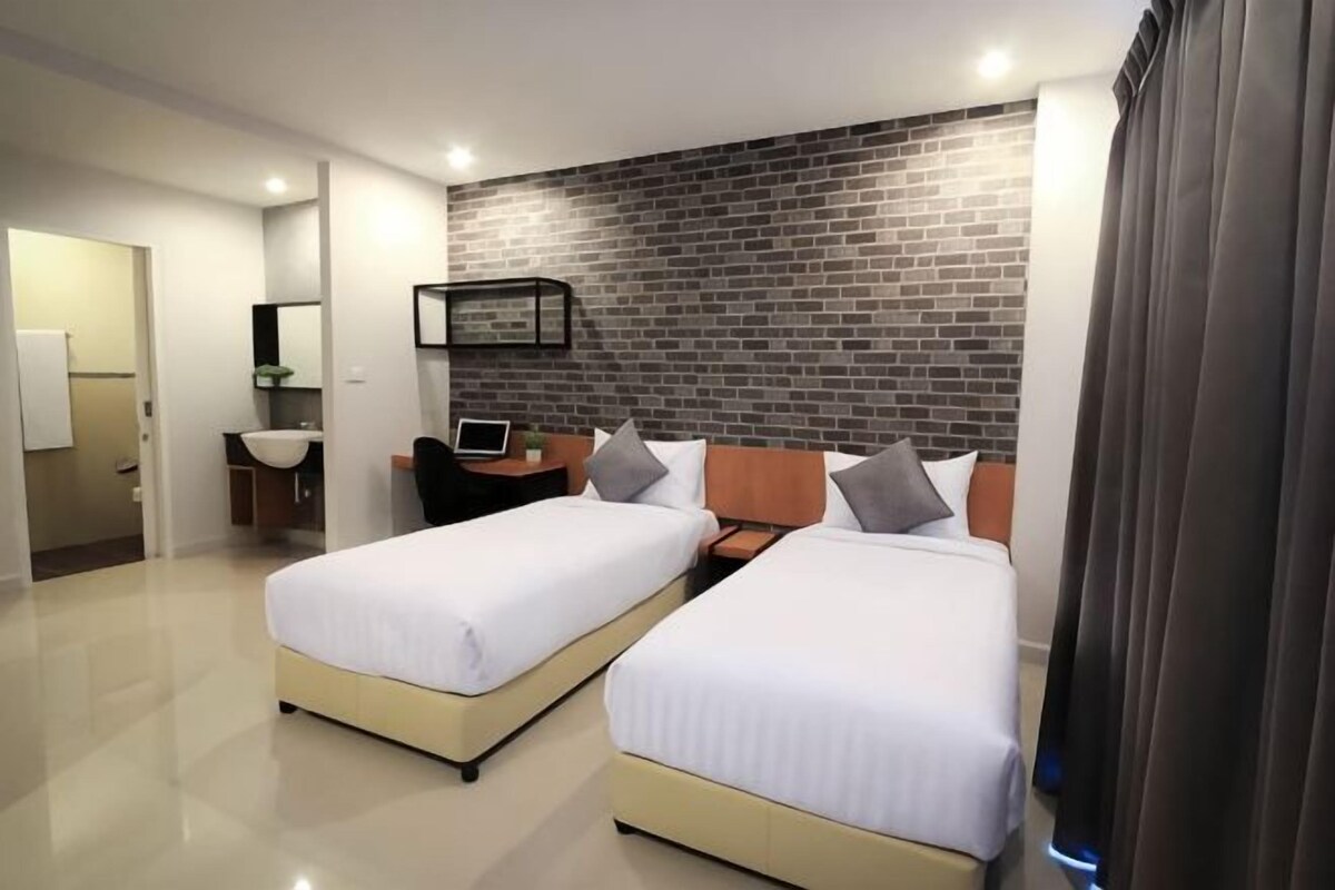 A hotel room with two beds, a desk, a chair, a laptop, a small sink, and a brick-patterned wall.