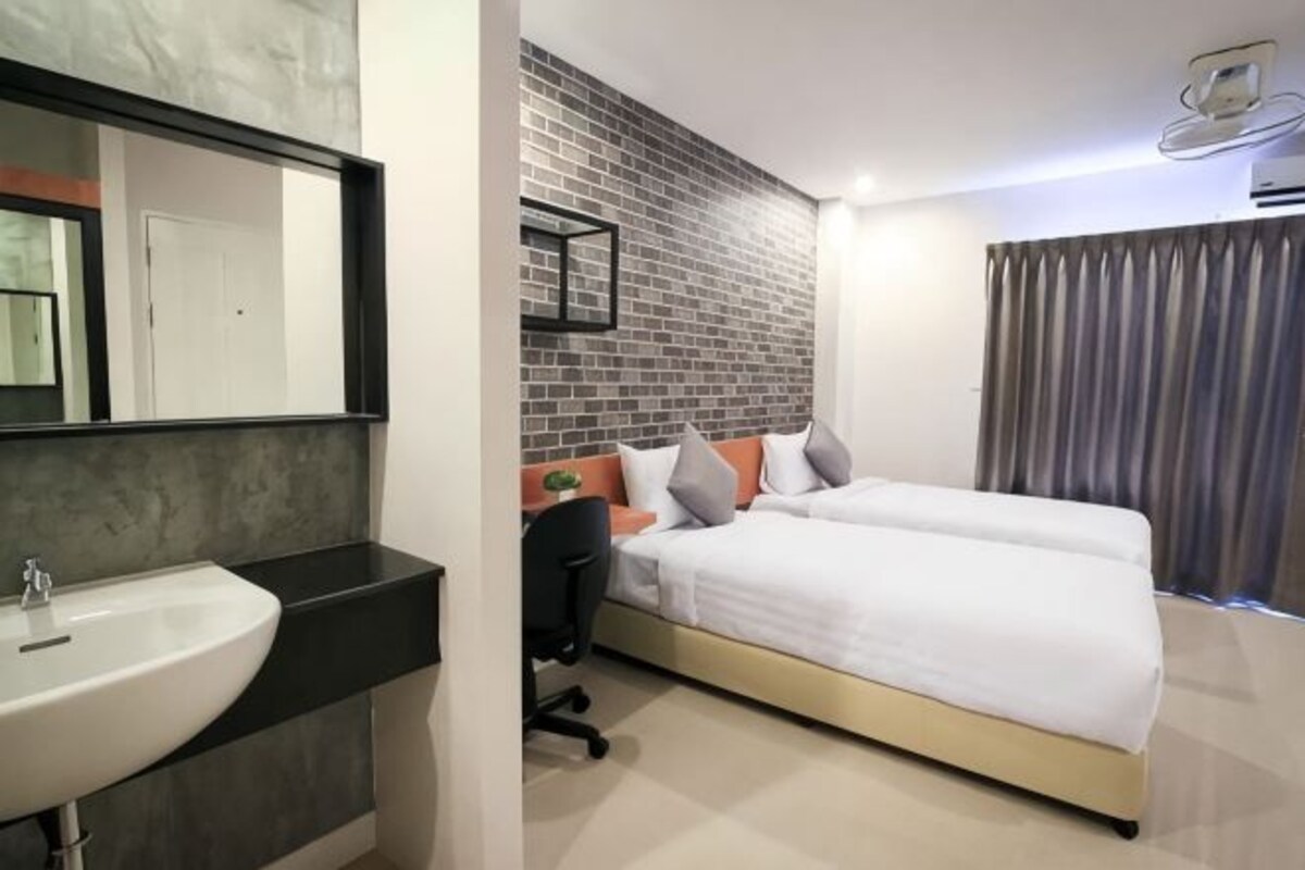 A modern hotel room with a large bed, a bedside table, a chair, a large mirror, and a bathroom with a sink.