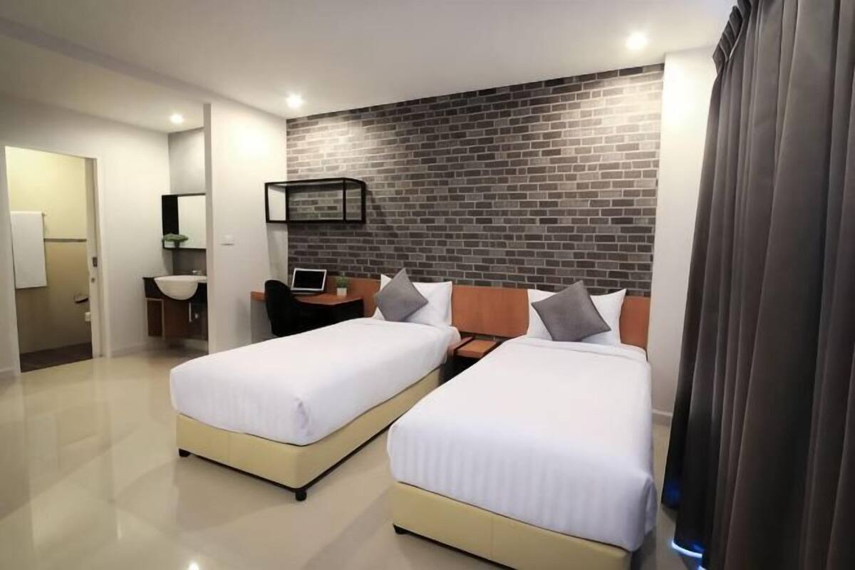 A hotel room with two beds, a desk, a chair, a laptop, a small sink, and a brick-patterned wall.
