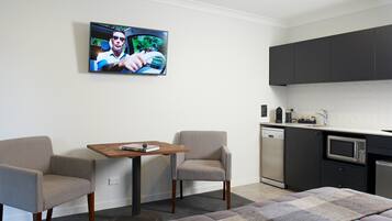 Una habitación moderna con un televisor de pantalla plana en la pared, una pequeña mesa de madera, dos sillones, un sofá, una zona de cocina con microondas y lavavajillas, y una alfombra estampada.