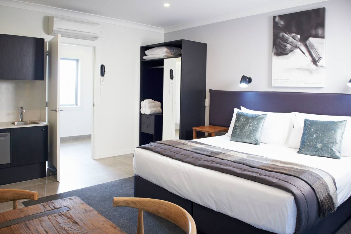 A modern hotel room with a large bed, a wooden dining table, a kitchen area, and a bathroom.