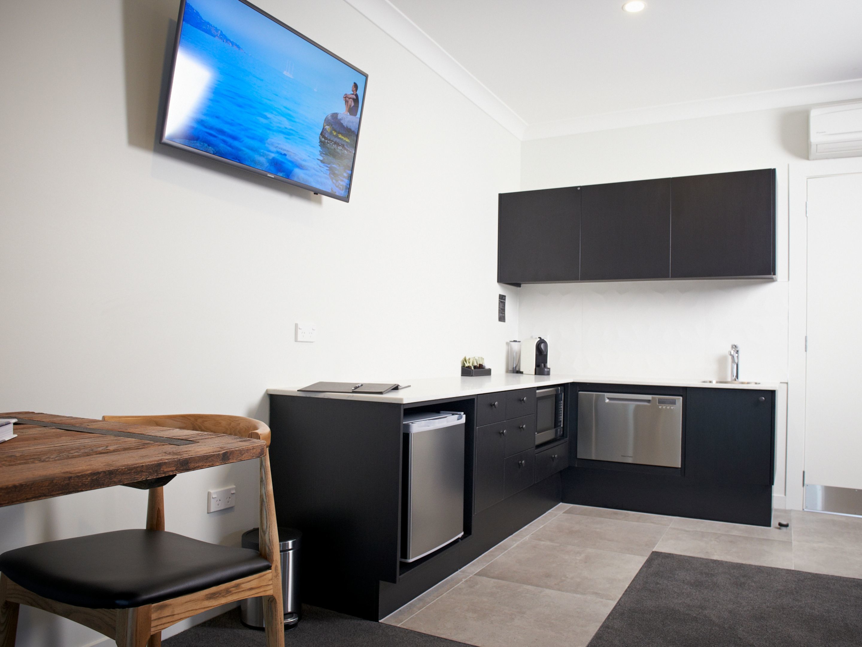 A modern kitchen with a wooden dining table and chairs, a built-in dishwasher, and a flat-screen TV mounted on the wall.