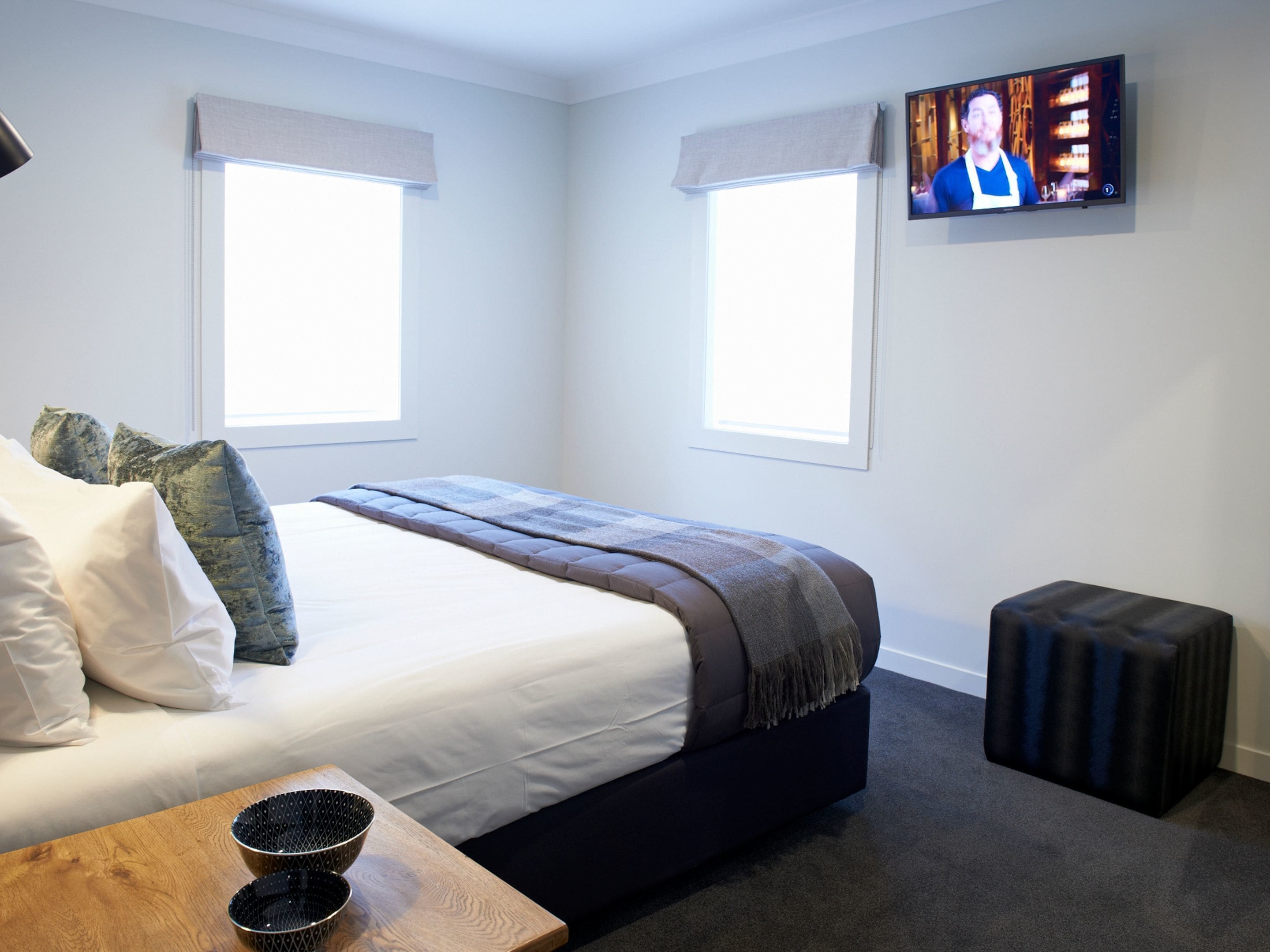 A modern hotel room with a large bed, two windows, a television on the wall, and a small wooden table with bowls.