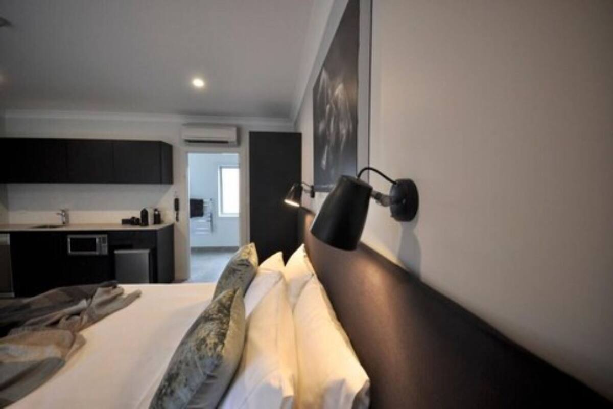 A modern hotel room with a bed, a bedside table, a wall-mounted lamp, and a view of the kitchen area.