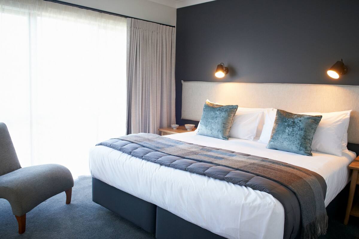A modern hotel room with a large bed, a chair, a nightstand, and a lamp.