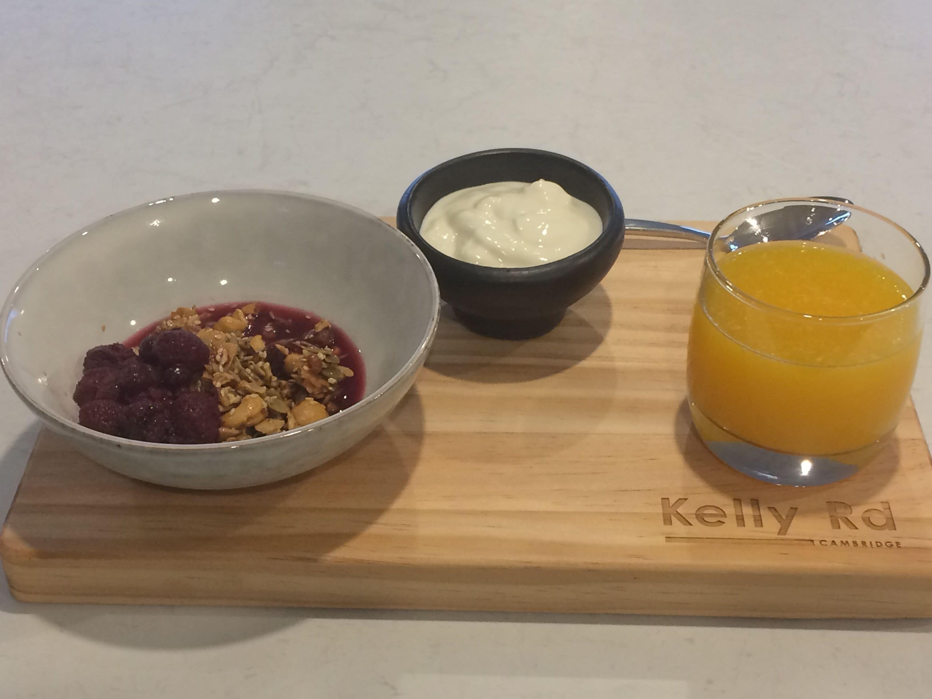 A breakfast tray with a bowl of fruit salad, a small bowl of yogurt, and a glass of orange juice on a wooden board with "Kelly Rd Cambridge" engraved on it.