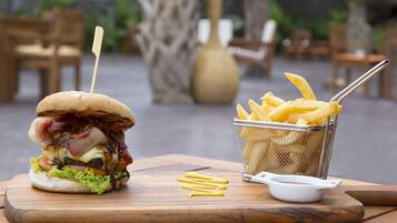 A burger with bacon, cheese, and lettuce on a wooden board, accompanied by a basket of French fries and a small white dish.