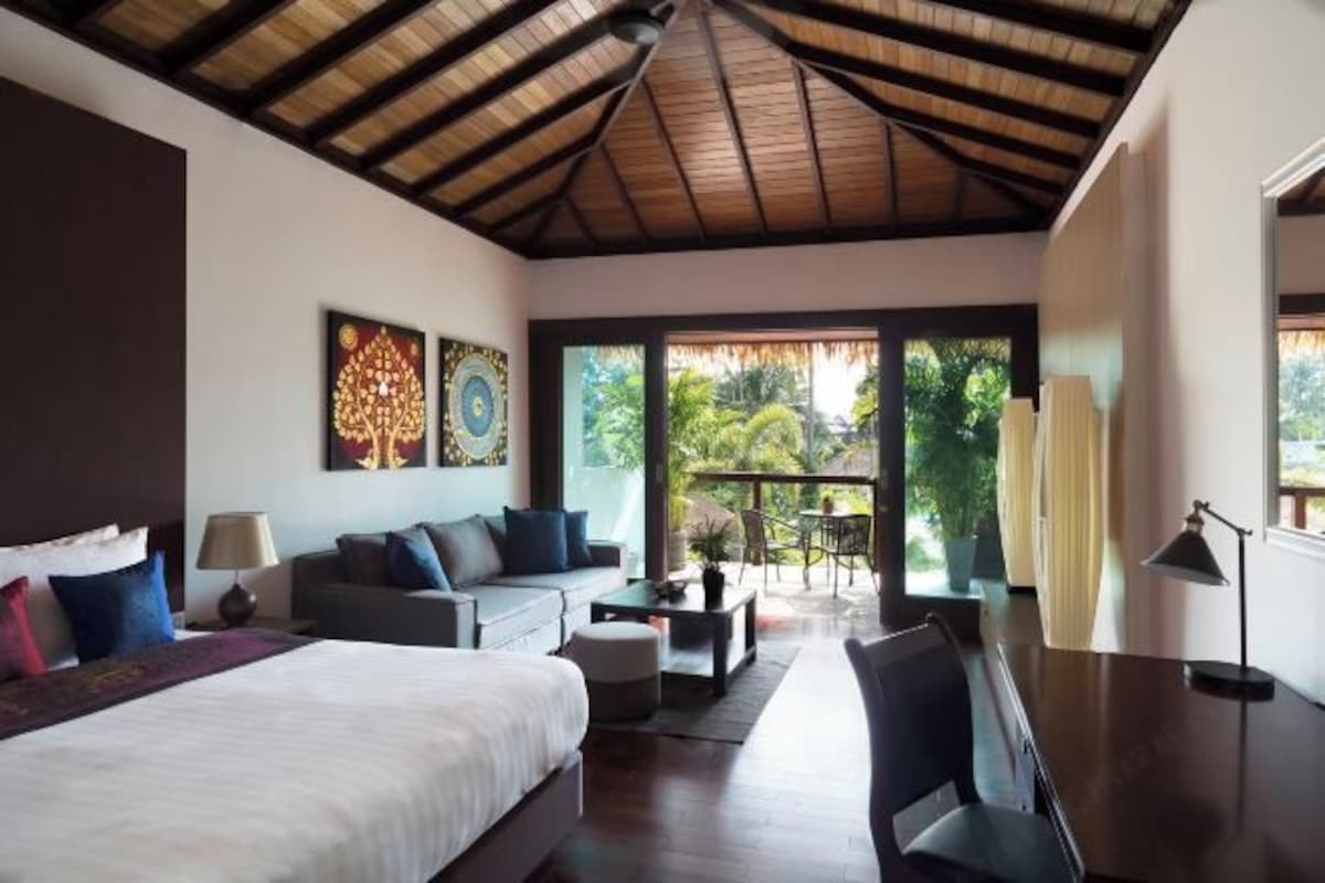 Habitación de hotel moderna con una cama grande, un sofá, una mesa de comedor y un balcón con vistas a zonas verdes.
