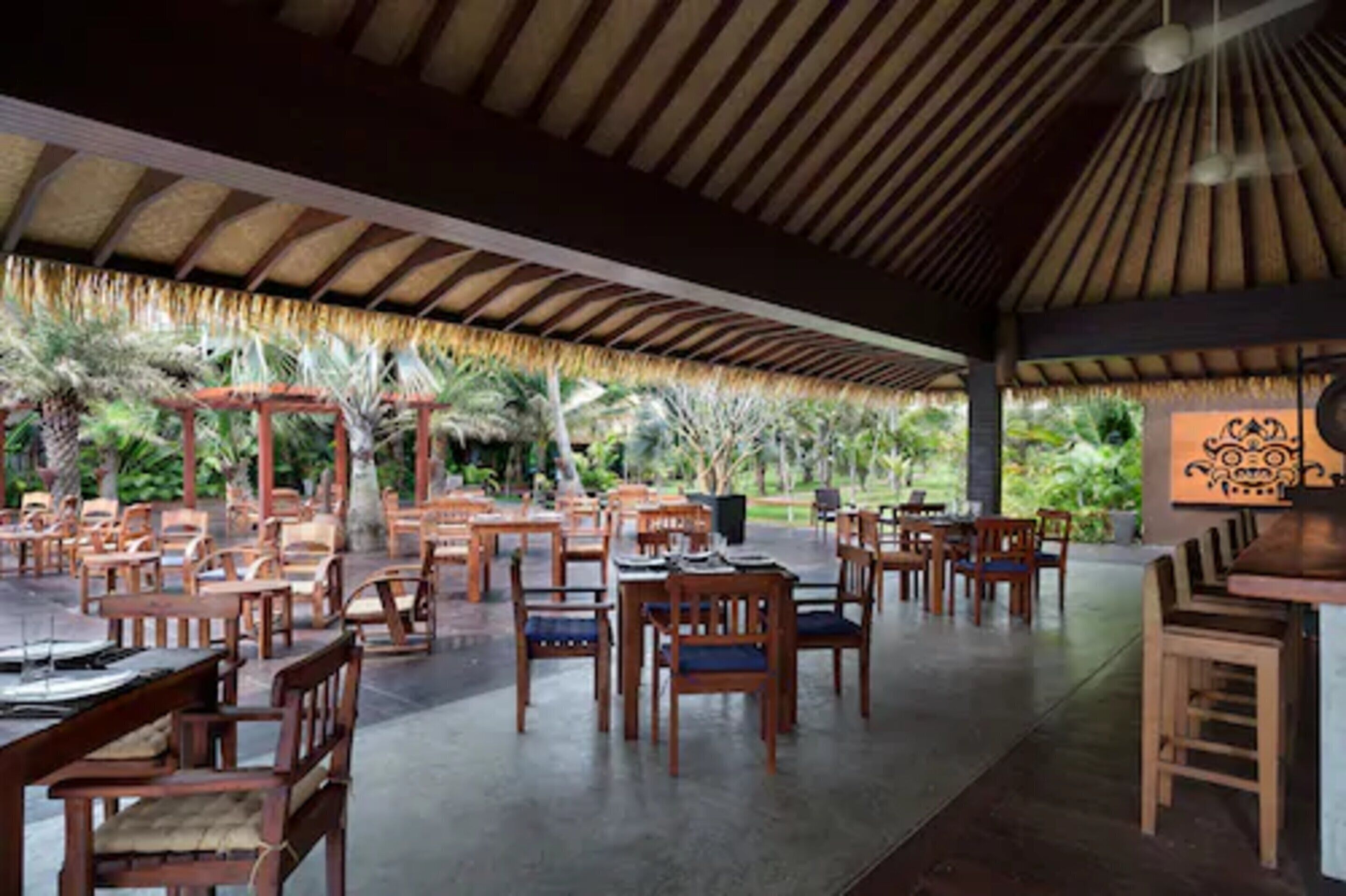 An outdoor dining area with wooden tables and chairs, surrounded by tropical plants and a thatched roof structure.