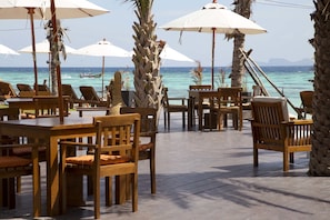An outdoor seating area with wooden tables and chairs, surrounded by palm trees and umbrellas, overlooking a calm sea.